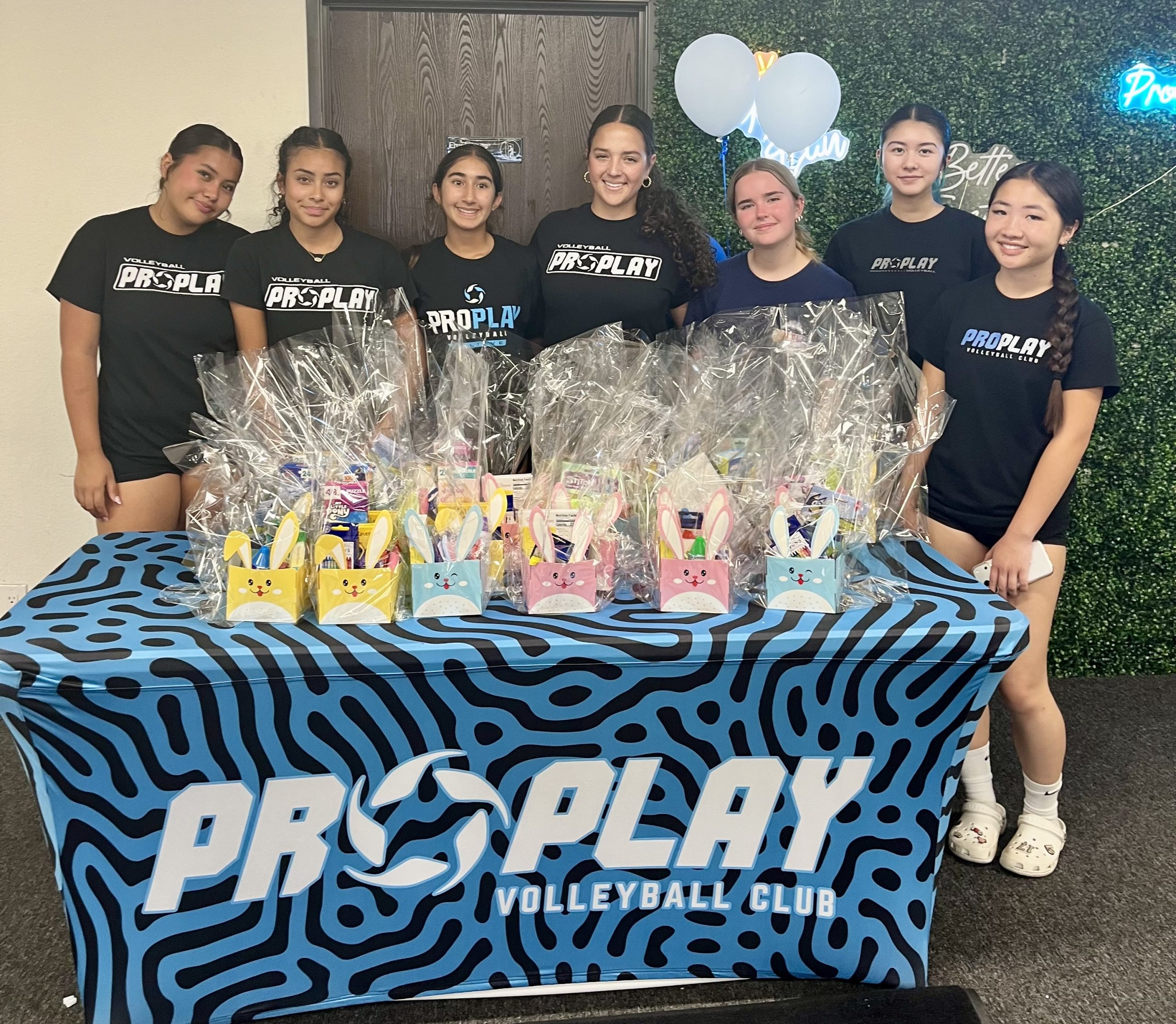 Seven ProPlay Volleyball team members hug together as they stand behind the Easter Baskets they made and smile.
